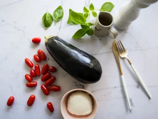 Bakłażan, pomidorki koktajlowe, ser mozzarella oraz listki bazylii leżące na kuchennym blacie