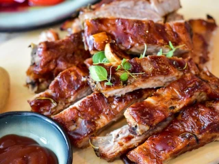 Upieczone żeberka BBQ pokrojone na porcje, z widoczną błyszczącą glazurą i świeżymi ziołami. Obok miseczka z sosem barbecue i ziemniak