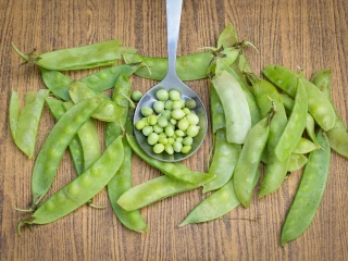 Świeże zielone strąki grochu rozłożone na drewnianym blacie, pośrodku metalowa łyżka wypełniona wyłuskanymi ziarnami groszku