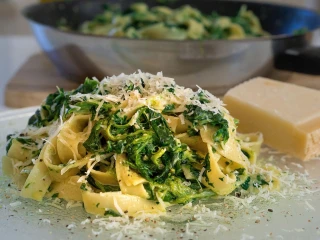 Porcja makaronu tagliatelle z kurczakiem i szpinakiem w kremowym sosie śmietanowym, posypana tartym serem na przezroczystym talerzu, w tle miska z resztą dania i kawałek sera