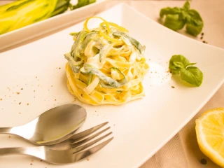 Elegancko uformowana porcja makaronu tagliatelle w sosie śmietanowym z zielonymi warzywami na dużym białym talerzu, podana z listkami bazylii, plasterkiem cytryny i kompletem sztućców