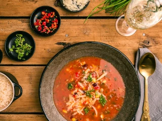 Miska zupy meksykańskiej z kukurydzą, pomidorami i serem na drewnianym stole, otoczona dodatkami jak ryż, papryczki chili, zioła, masło i kieliszek białego wina