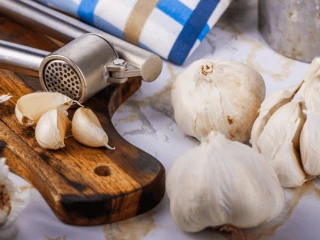 Na drewnianej desce leżą ząbki czosnku oraz praska do czosnku