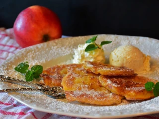 Placki z jabłkami oprószone cukrem pudrem, podane na talerzu z gałązką mięty, bitą śmietaną i gałką lodów waniliowych, w tle czerwone jabłko