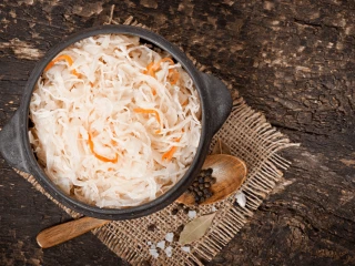 Kapusta kiszona w szarej misce na rustykalnej ścierce, obok drewniana łyżka z ziarnami pieprzu