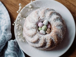 Babka majonezowa udekorowana cukrem pudrem, pisankami i gałązkami z białymi pąkami na białym talerzu