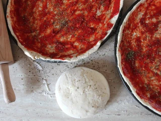 Surowe ciasto na pizzę wyłożone na dwóch blachach i posmarowane pomidorowym sosem, obok leży kulka świeżego ciasta i drewniany wałek