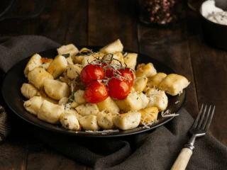 Czarny talerz usmażonych kopytek z gałązką pomidorków koktajlowych. całość posypana parmezanem