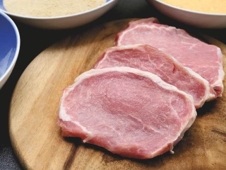 Trzy kawałki surowego schabu leżą na drewnianej desce, w tle trzy miski ze składnikami panierki