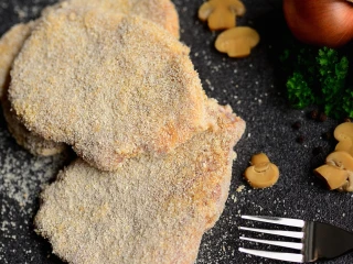 Na czarnym blacie leżą trzy surowe, zapanierowane kotlety schabowe, obok leży widelec i pieczarki