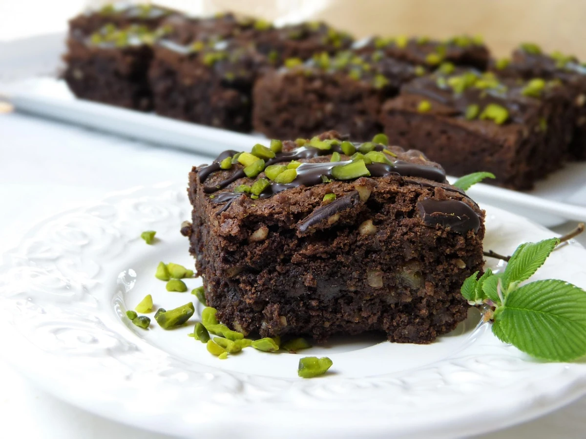 Kawałek czekoladowego brownie ozdobionego kawałkami pistacji i polewą czekoladową, podany na eleganckim białym talerzu