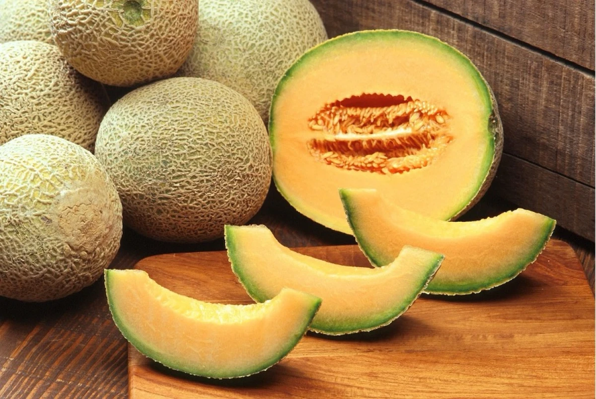 Na drewnianej desce leży pokrojony na kawałki melon kantalupa, w tle całe owoce