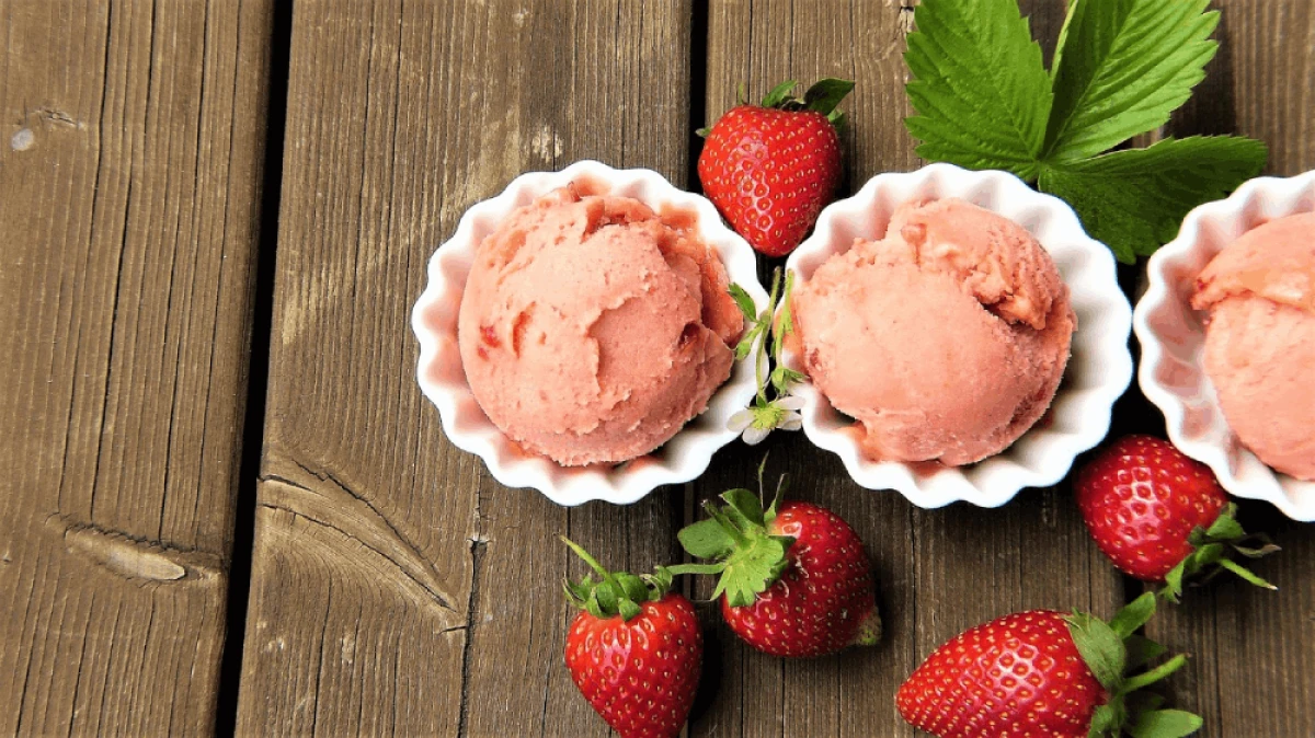 Na drewnianym blacie leżą trzy białe miseczki z trzema gałkami lodów truskawkowych, obok leżą truskawki