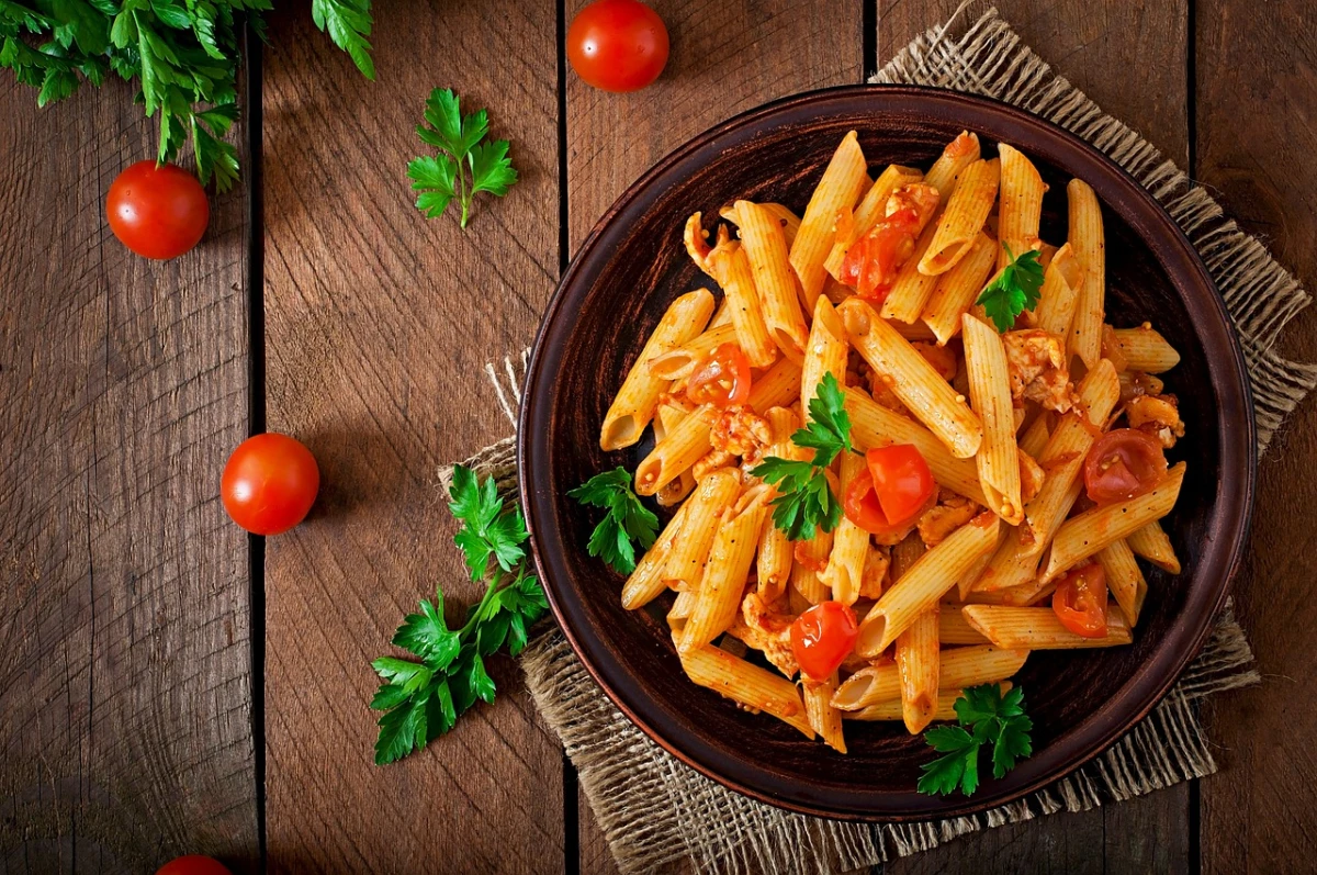 Na drewnianym blacie stoi brązowy talerz z makaronem penne i pomidorami. 