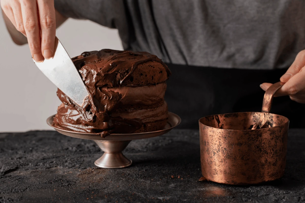 Osoba ozdabia czekoladowy tort czekoladową polewą, obok miedziany garnek z czekoladą