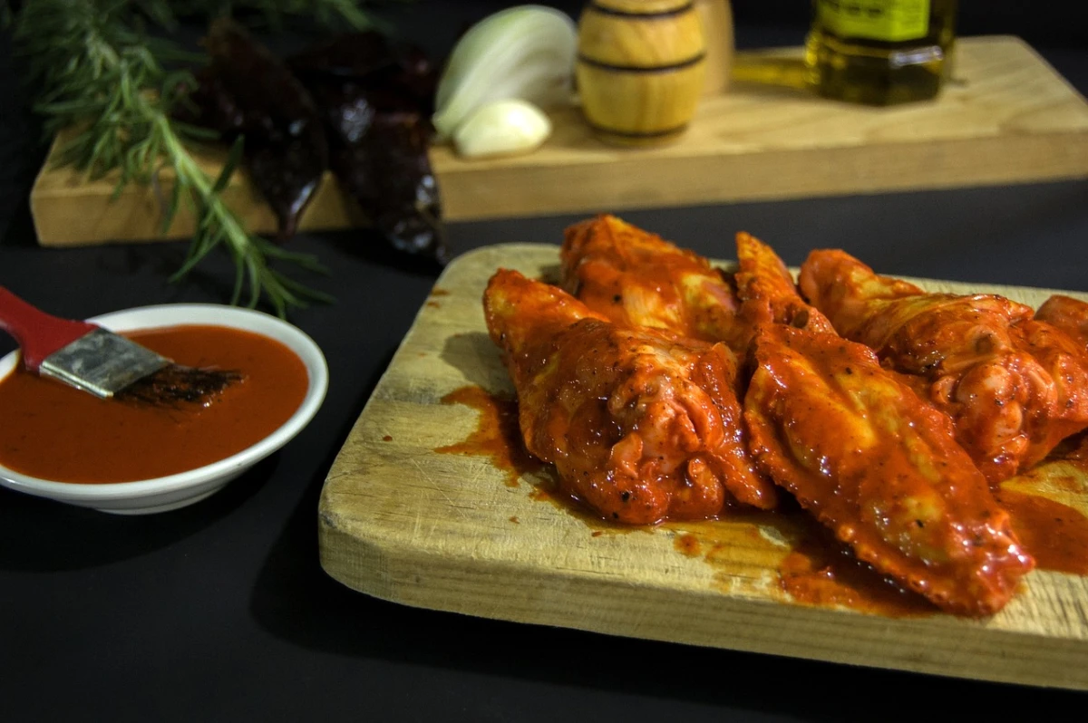 Soczyste kawałki kurczaka w czerwonej marynacie, leżą na drewnianej desce, obok miska z marynatą i pędzelek