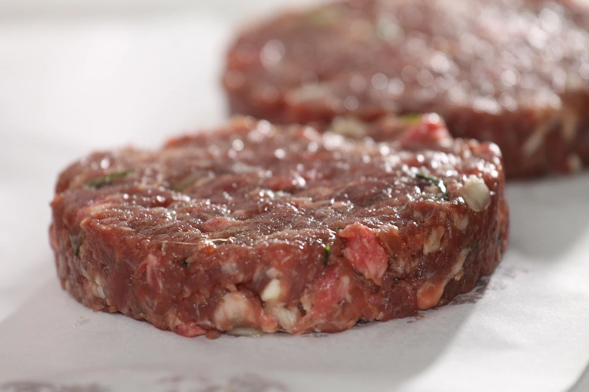 Surowy, uformowany kotlet z mielonej wołowiny z widocznymi kawałkami tłuszczu i przypraw, leżący na papierze do pieczenia