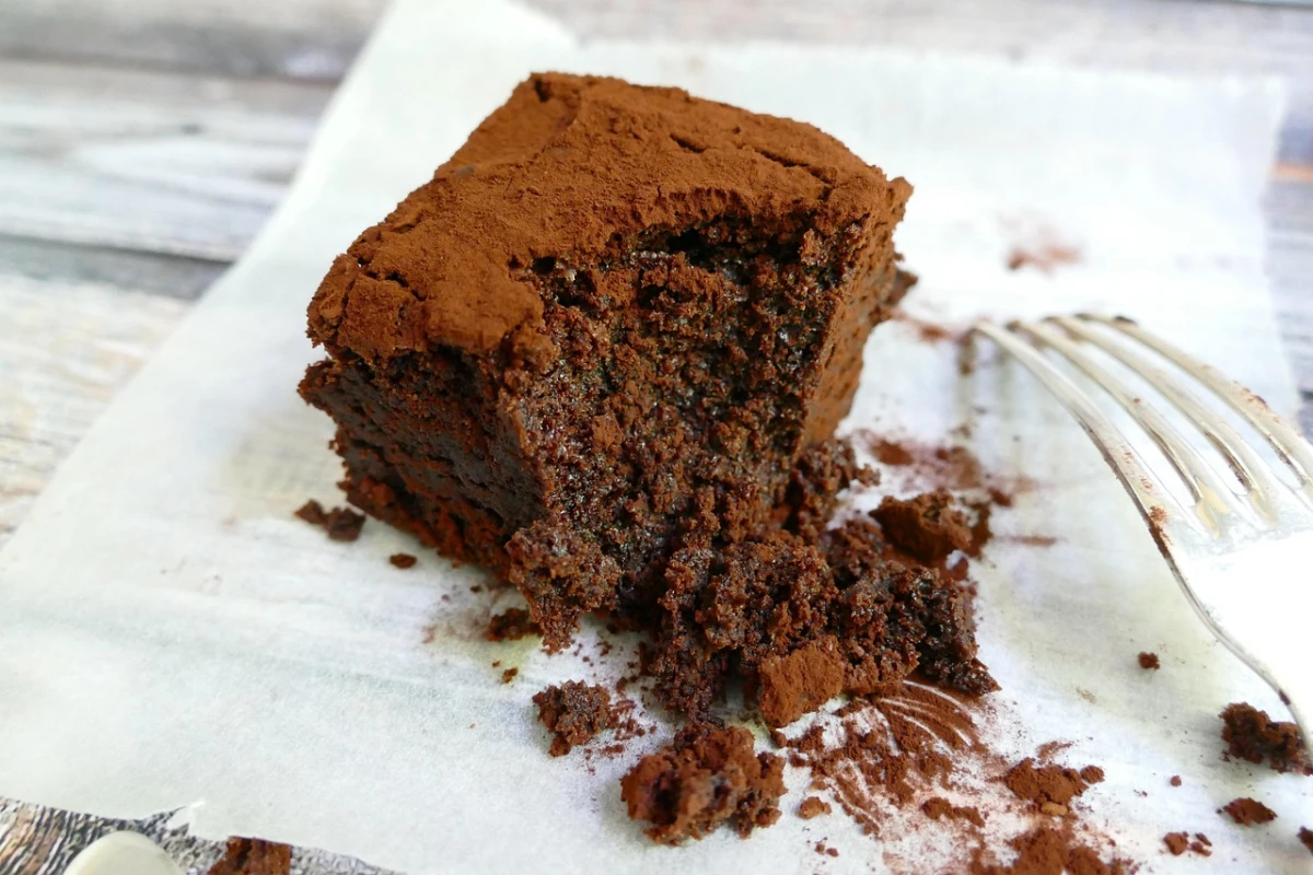 Wilgotny kawałek brownie, obsypany warstwą kakao, z widocznym ugryzieniem, na papierze do pieczenia obok widelca