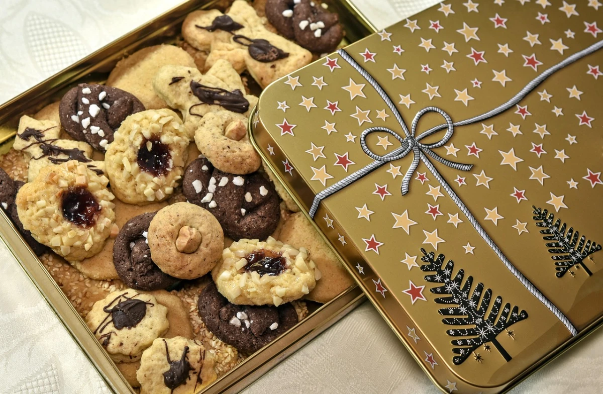 Złota puszka świąteczna, wypełniona różnorodnymi domowymi ciasteczkami - z marmoladą, kakao, polewą czekoladową i posypką, ułożone obok siebie na jasnym obrusie